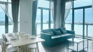 A modern room with a dining table set for four, a sofa, and large windows offering a view of the sea. - Kwen Suites - City Centre Riverson Soho 2Br l Studio l Duplex (Kota Kinabalu)