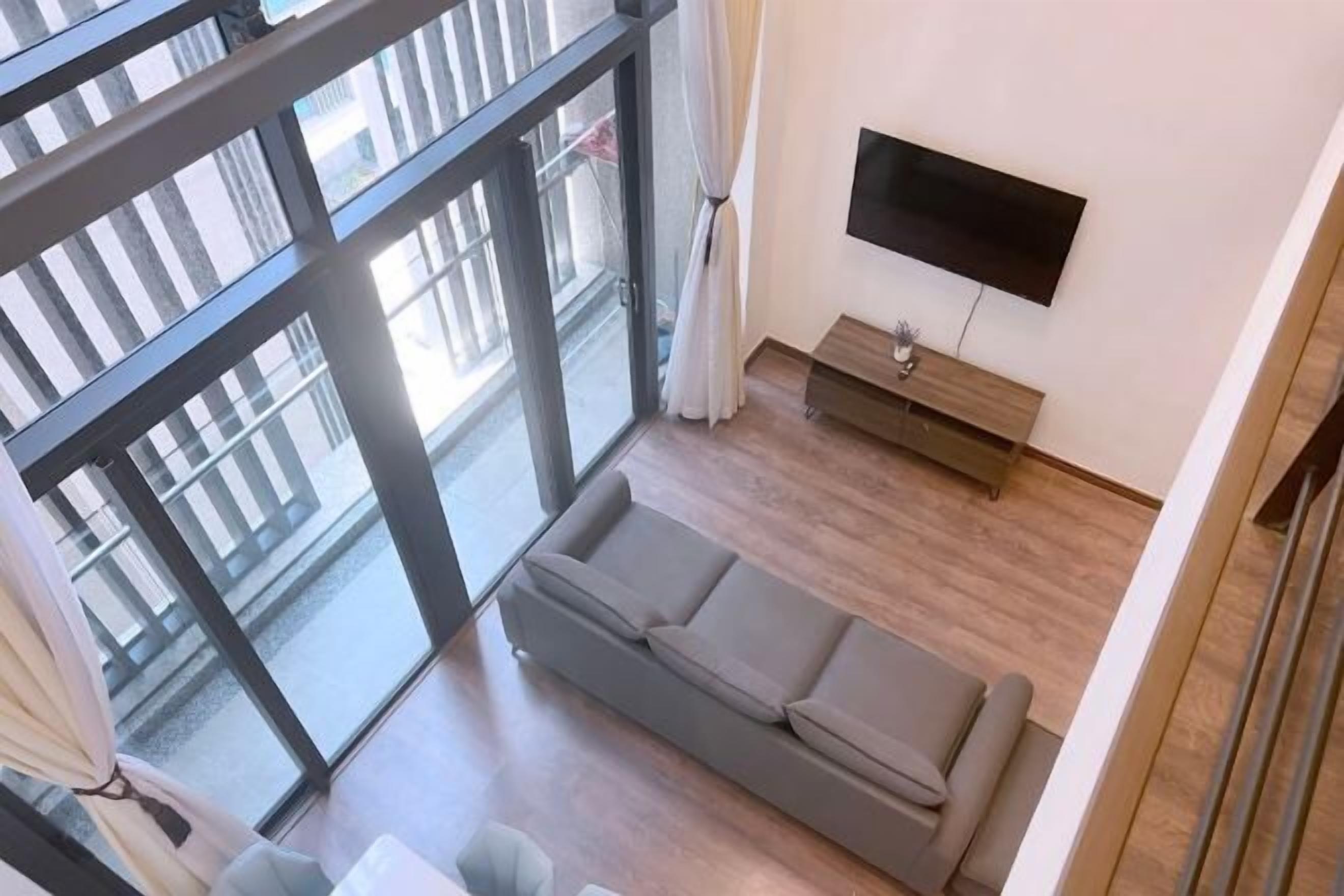 A modern living room with a grey sofa, a flat-screen TV mounted on the wall, and a sliding glass door leading to an outdoor balcony.