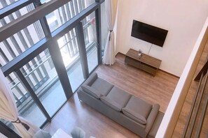 Un salon moderne avec un canapé gris, un téléviseur à écran plat fixé au mur et une porte-fenêtre coulissante donnant sur un balcon extérieur.