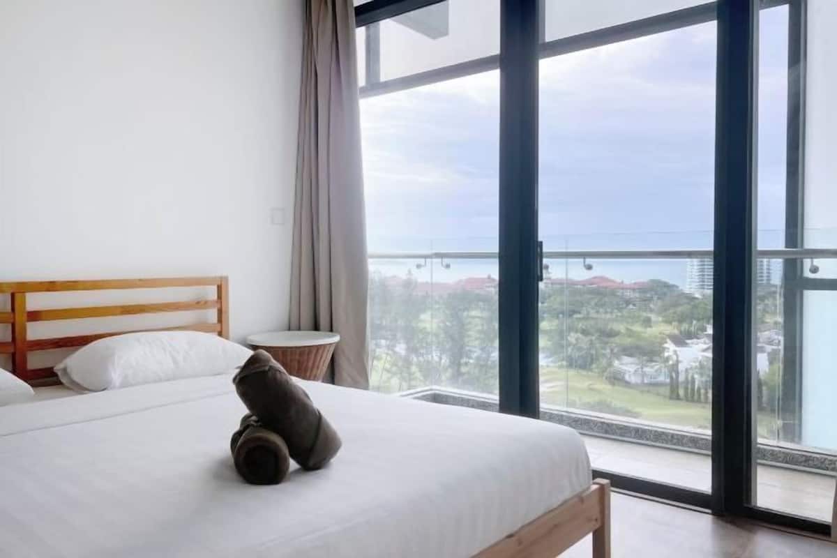 Ein Schlafzimmer mit einem Holz-Kopfteil, einem Bett mit weißer Bettwäsche, einem runden Beistelltisch und Blick auf eine Stadtlandschaft durch große Fenster.