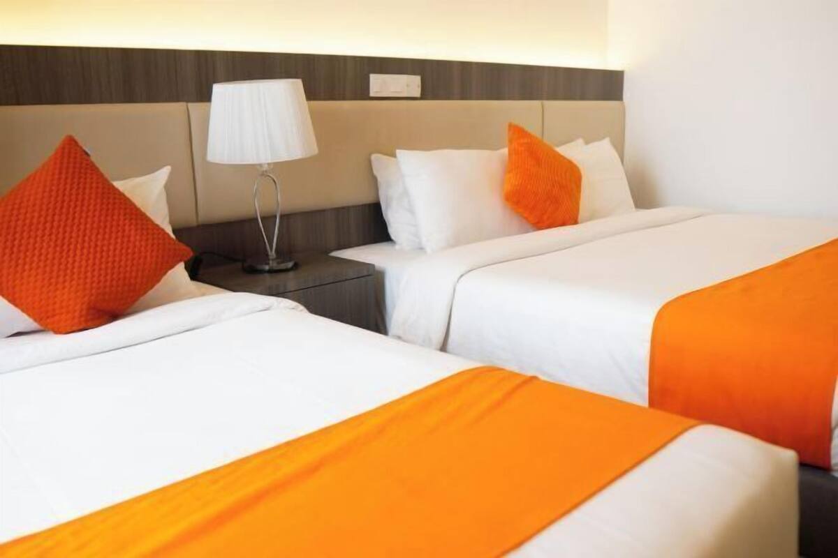 Camera d'albergo con due letti, entrambi con biancheria bianca e dettagli arancioni, un comodino con una lampada e una testiera con interruttore della luce.