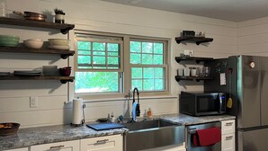 A modern kitchen with white cabinets, a stainless steel sink, a microwave, and a refrigerator. There are windows providing natural light, and a wooden floor. - Snuggle up and relax with the whole family at this Ava home.  (Ava)