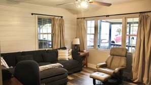 A living room with a dark-colored sofa, a recliner, a coffee table, and a ceiling fan. There are windows with curtains, a lamp on a side table, and a view of the outside. - Snuggle up and relax with the whole family at this Ava home.  (Ava)