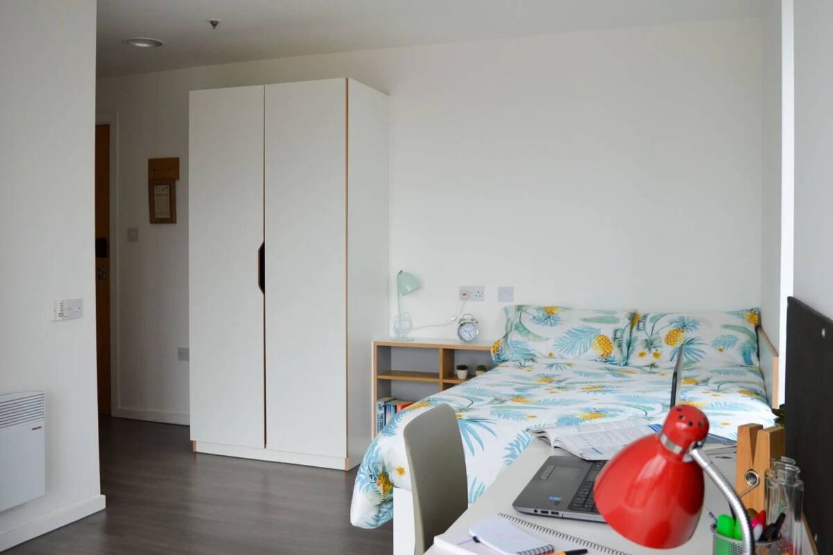 A bedroom with a bed, a desk, a laptop, a red lamp, and a wardrobe.