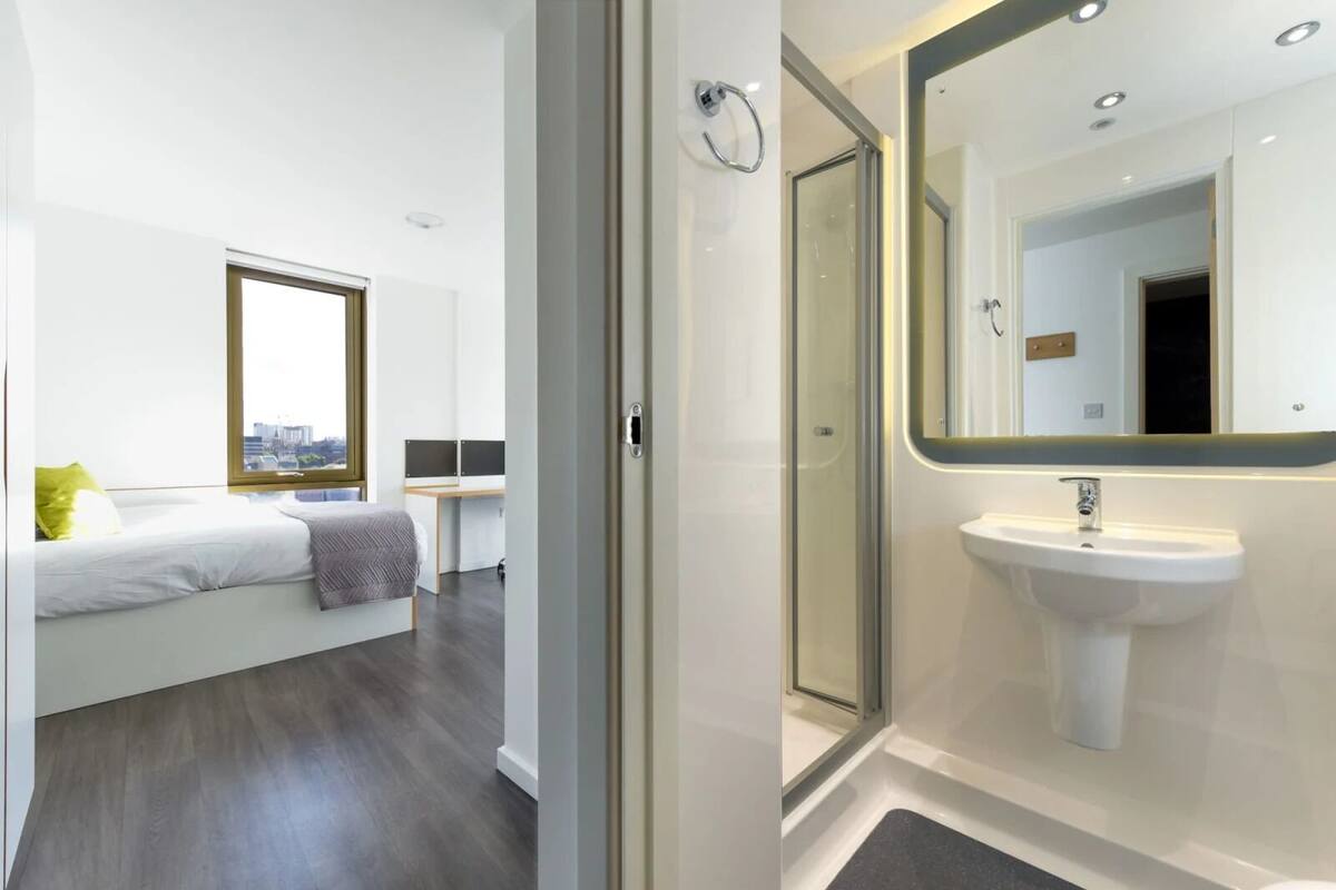 A modern bedroom with a bed, a desk, and a view of the city. It also features a bathroom with a sink and a glass shower enclosure.