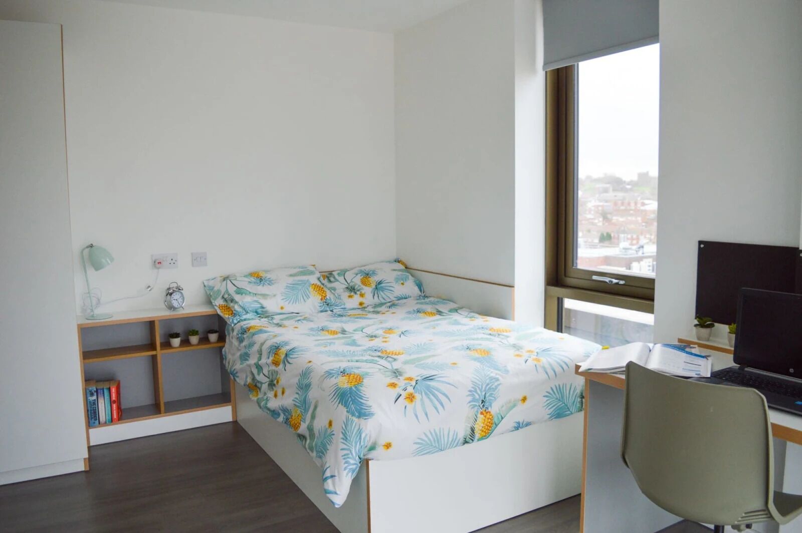 A small, modern bedroom with a bed, a desk with a computer, and a window with a city view.