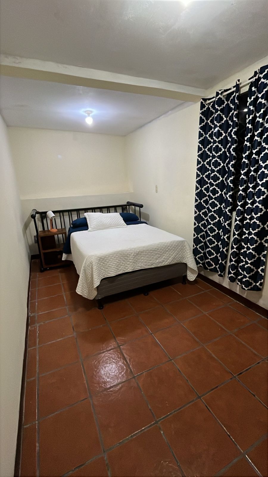 Una piccola camera con un letto singolo, pavimento piastrellato, un comodino, una lampada e una tenda decorata.