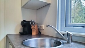 Et køkken med en stålvask, kaffemaskine og et vindue med udsigt til træer.