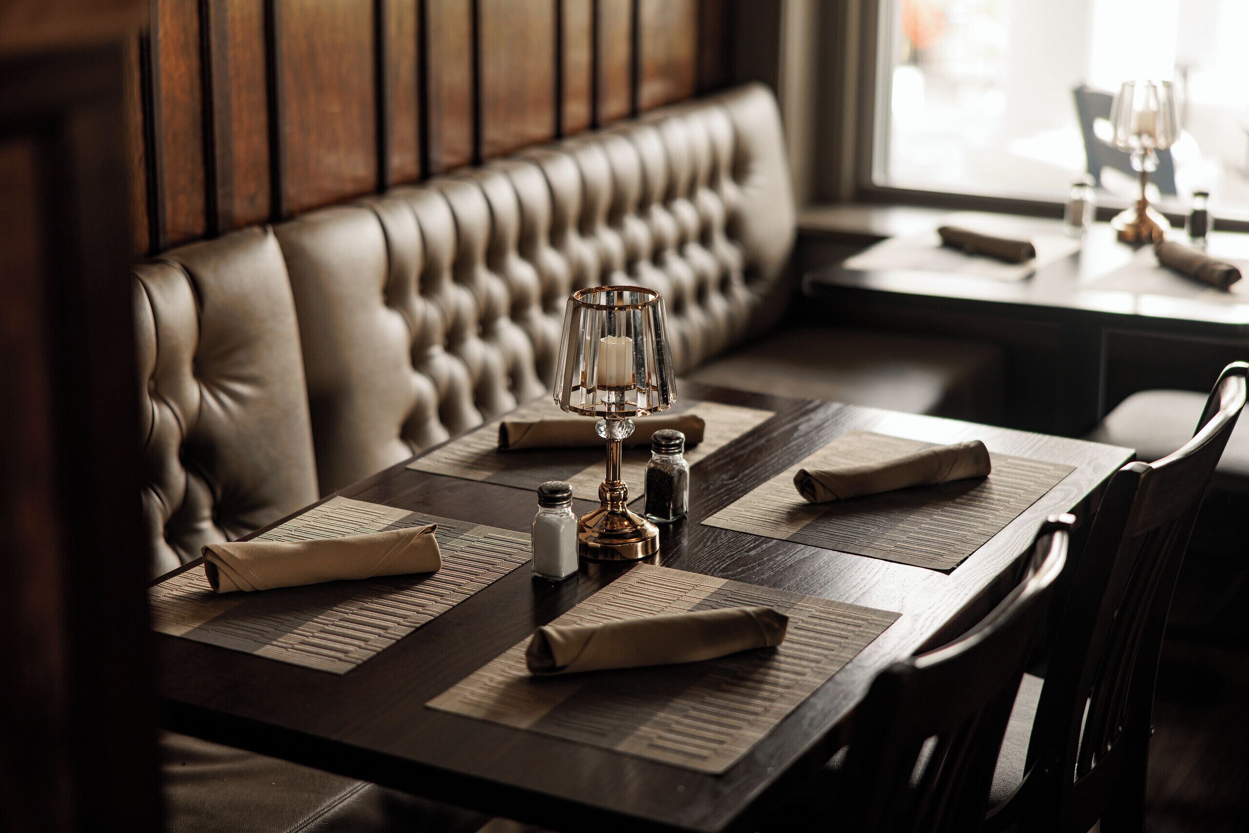 Een restaurantinterieur met een houten tafel gedekt voor twee personen, een gestoffeerde bank, zout- en pepervaatjes en een kandelaar.