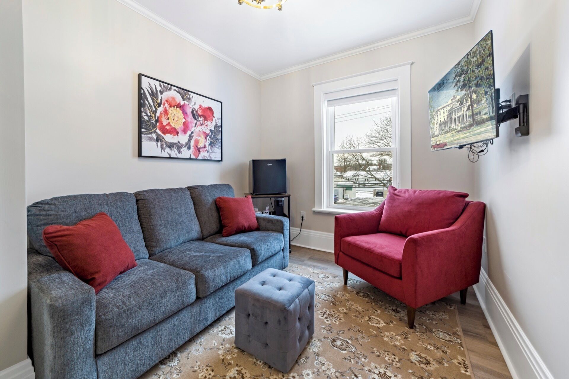 A living room with a grey sofa, red cushions, a red armchair, a grey ottoman, a flat-screen TV on a stand, and a window with a view of trees and a building.