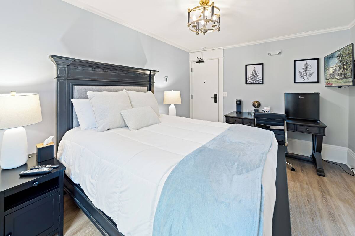 Ein Hotelzimmer mit einem großen Bett, Nachttischen mit Lampen, einem Schreibtisch mit Computer und gerahmten Bildern an der Wand.