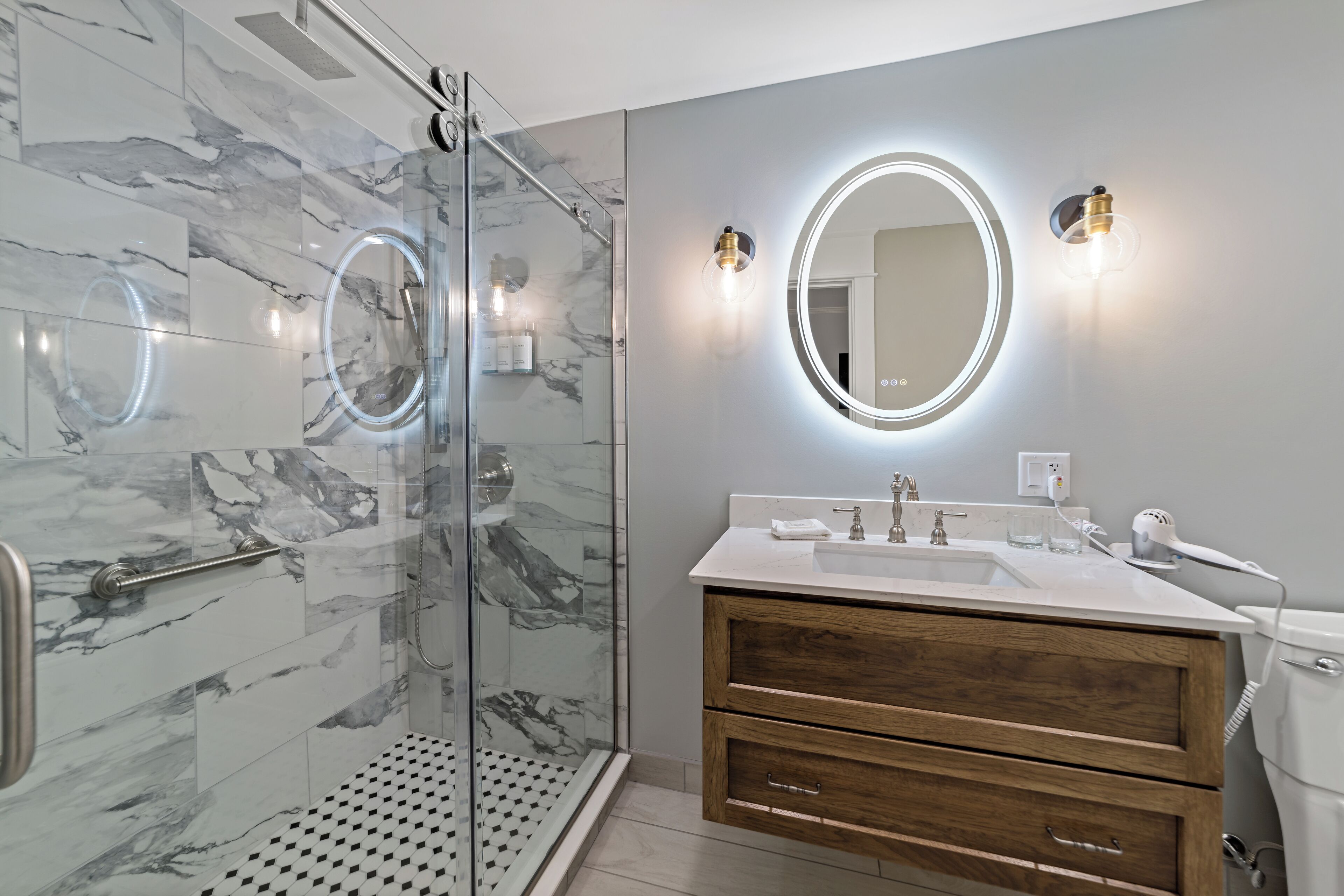 A modern bathroom with a glass-enclosed shower, marble tiles, a wooden vanity, and a round mirror with lighting.