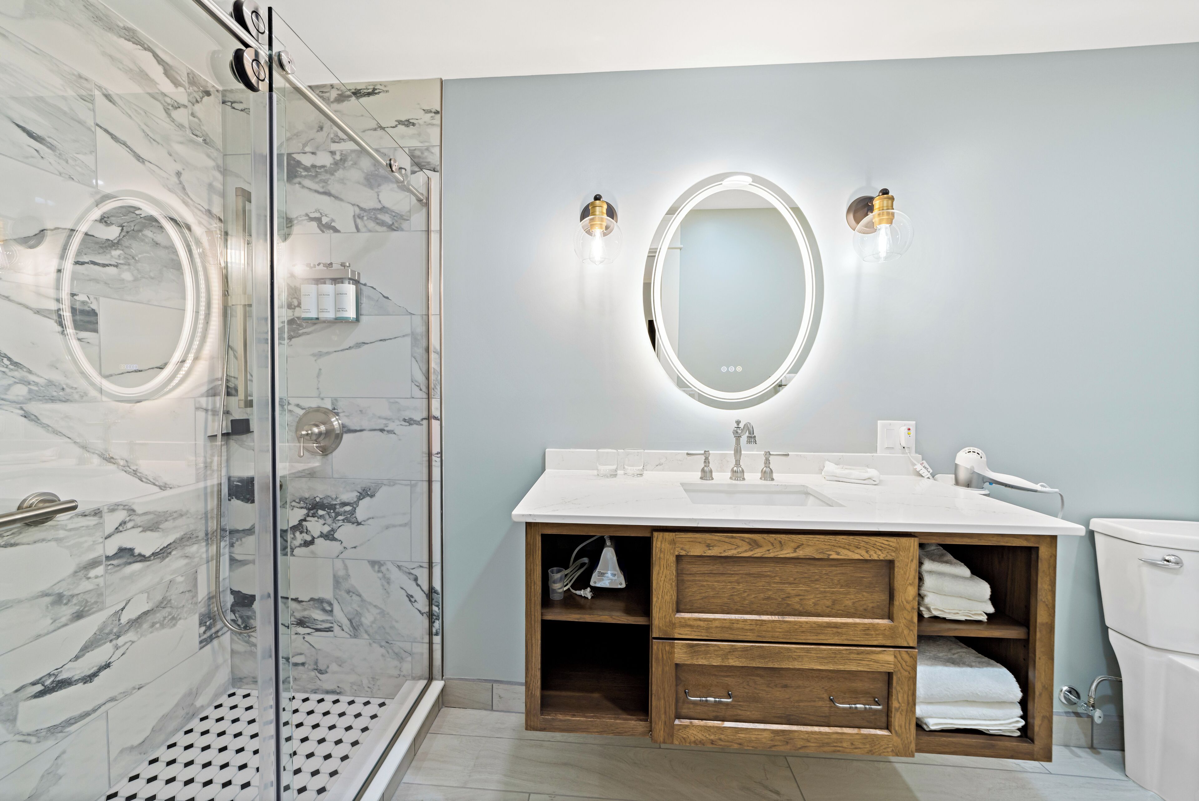 A modern bathroom with a glass-enclosed shower, a marble-tiled wall, a wooden vanity with a sink, and two wall-mounted lights.