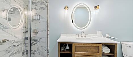 A modern bathroom with a glass-enclosed shower, a marble-tiled wall, a wooden vanity with a sink, and two wall-mounted lights.