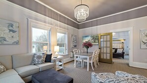 A spacious living room with a gray sofa, a dark ottoman, a dining table with chairs, and a chandelier. - The White Inn (Fredonia)