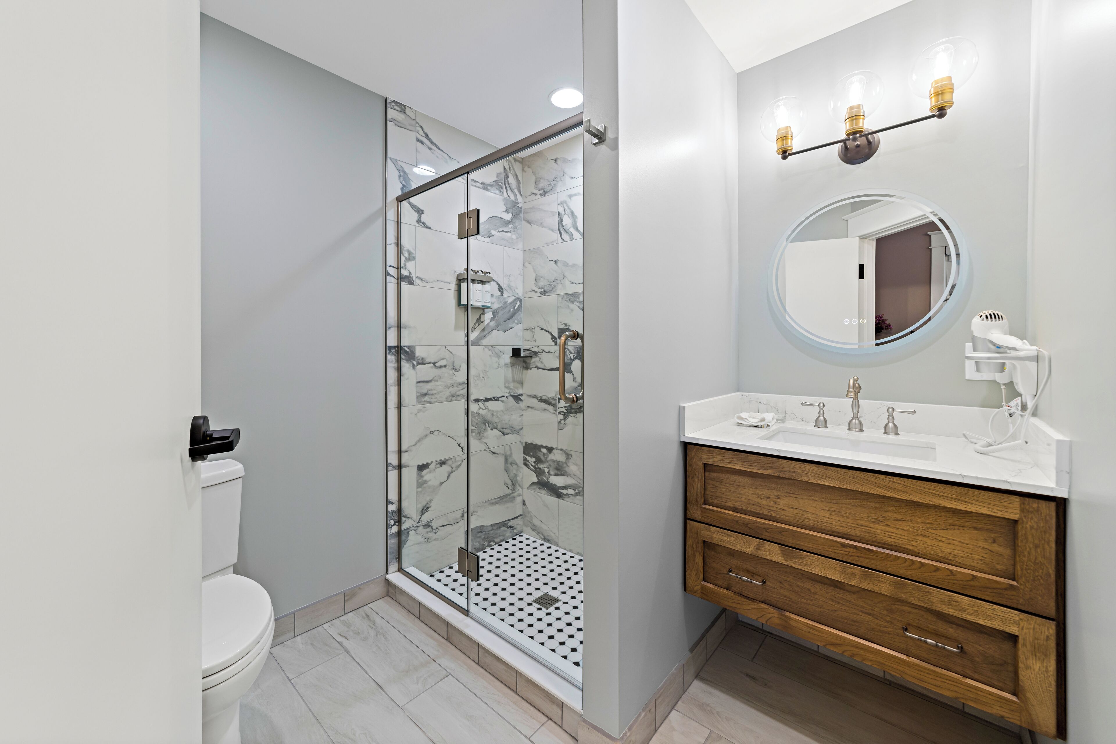 A modern bathroom with a glass-enclosed shower, a white toilet, a wooden vanity with a round mirror, and a white sink.
