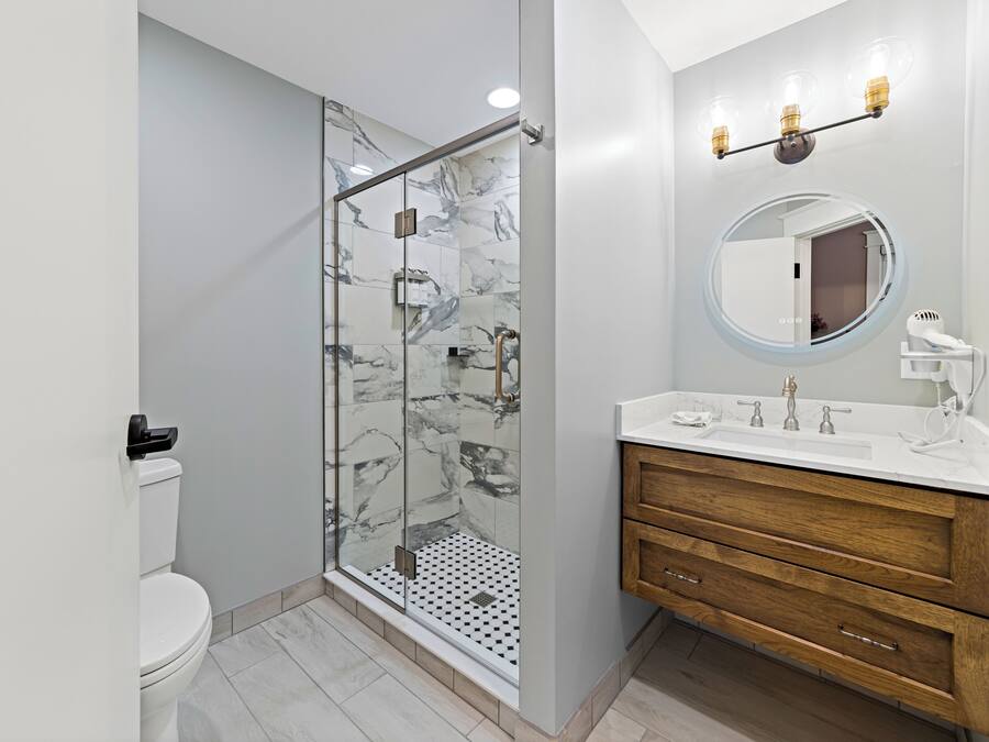 A modern bathroom with a glass-enclosed shower, a white toilet, a wooden vanity with a round mirror, and a white sink.