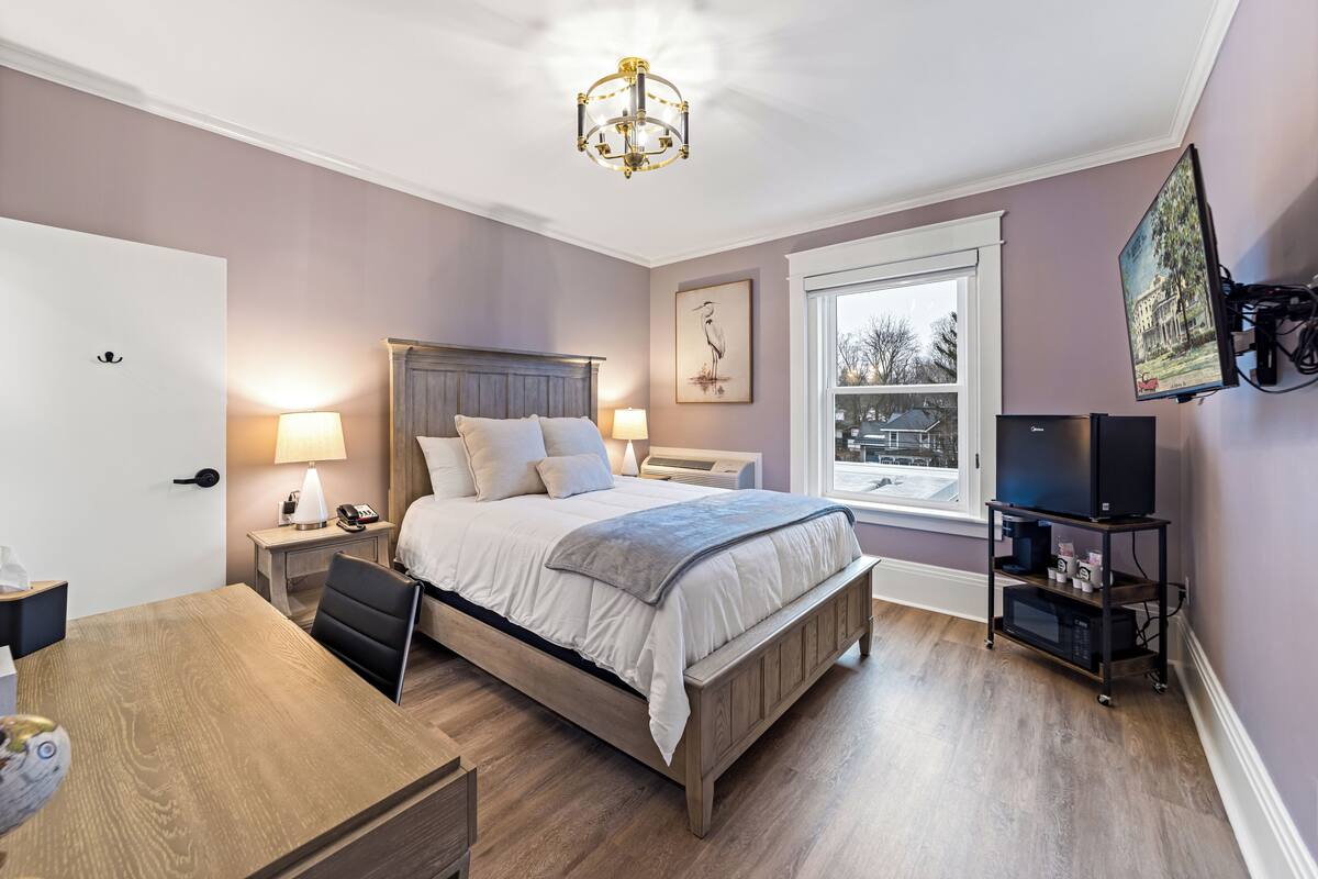 Ein Schlafzimmer mit Bett, Nachttischlampen, Schreibtisch, Stuhl, Fernseher auf einem Schrank und einem Fenster mit Blick.