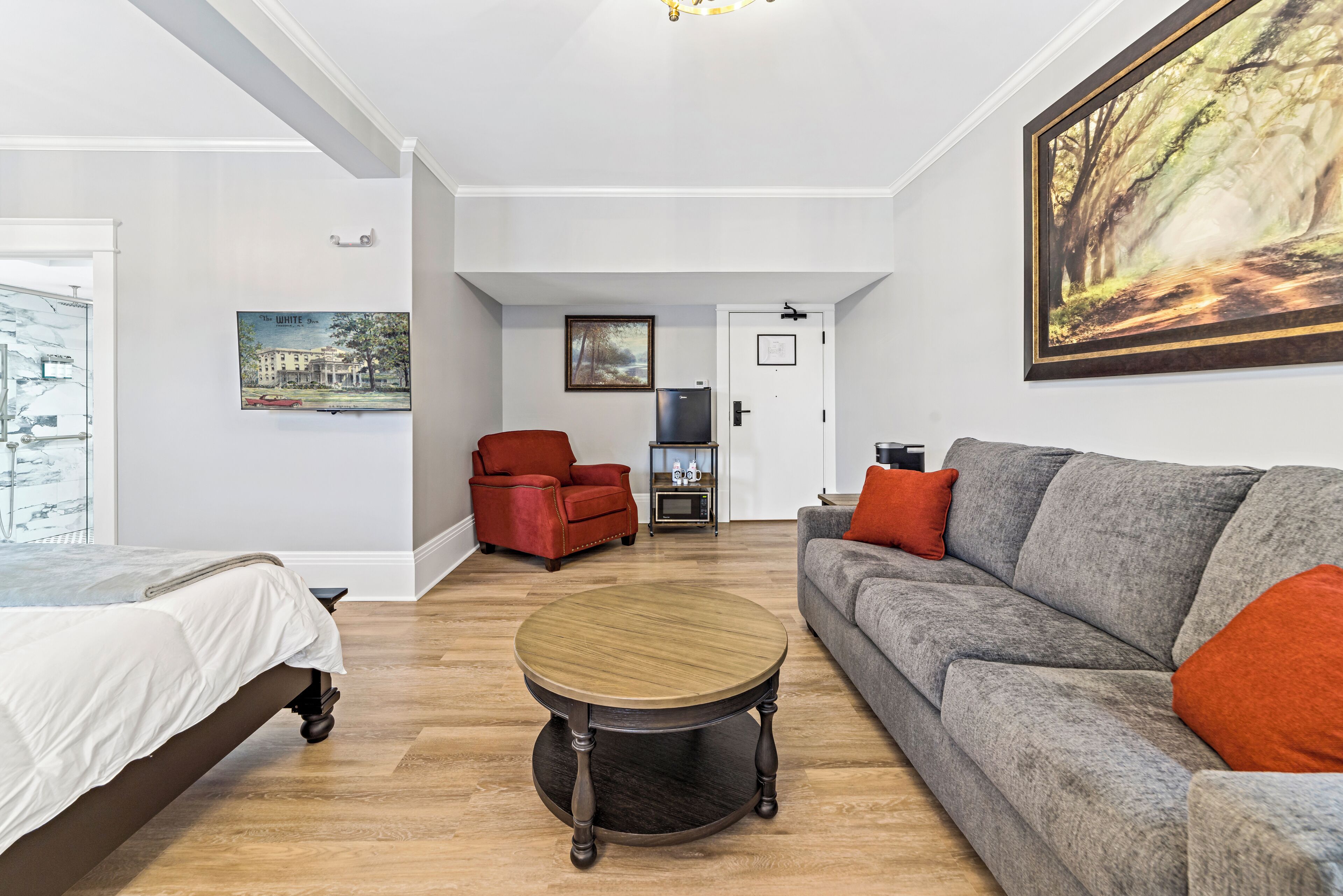 A modern living room with a bed, a grey sofa with orange cushions, a red armchair, and a round wooden coffee table.