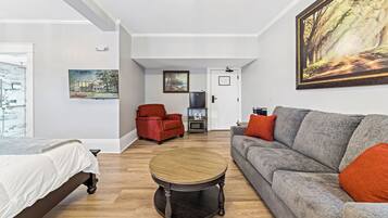 A modern living room with a bed, a grey sofa with orange cushions, a red armchair, and a round wooden coffee table.