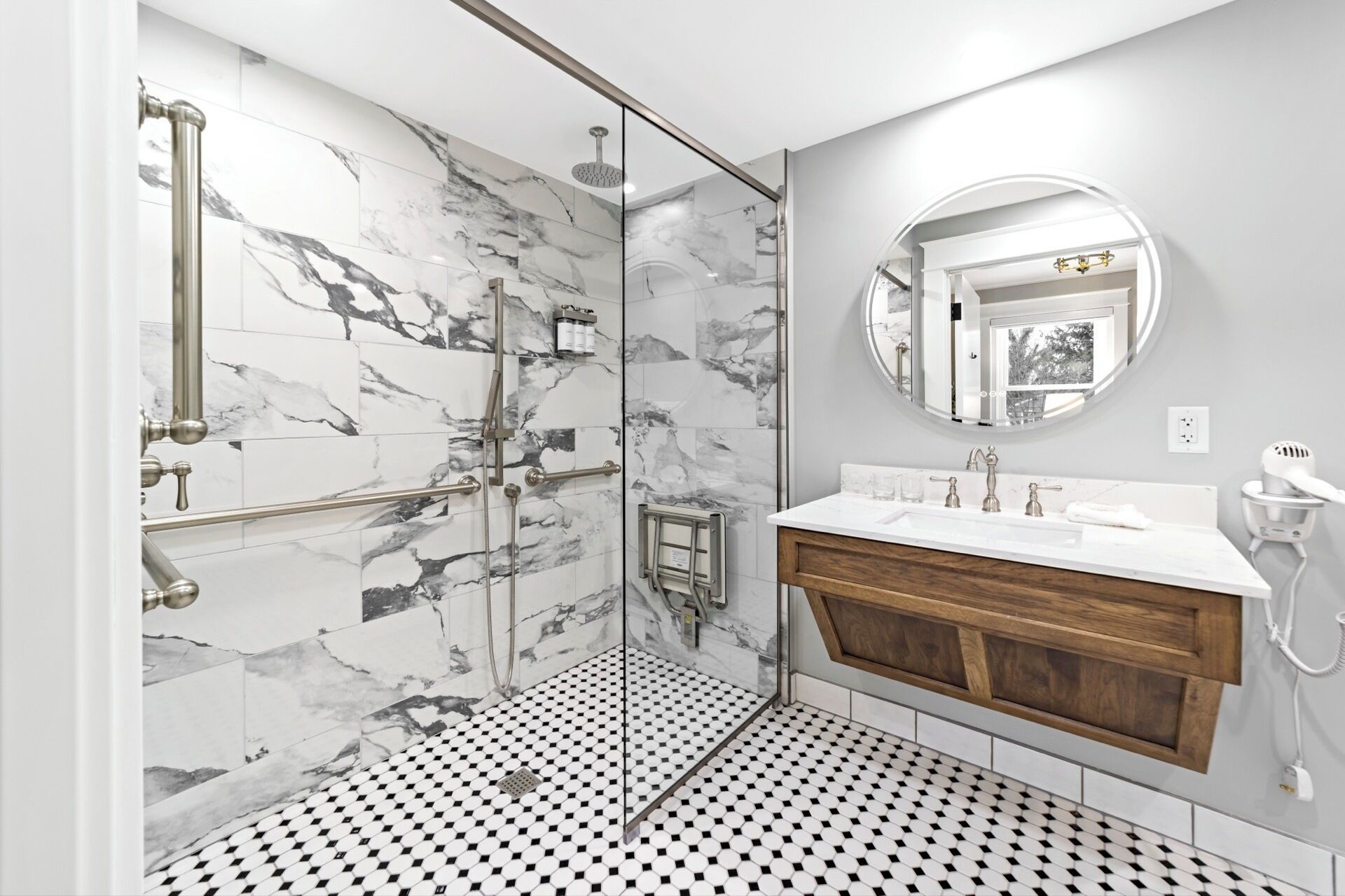 A modern bathroom with a shower, marble tiles, a wooden vanity, and a round mirror.