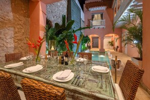 A dining area with a glass table set for four, surrounded by wicker chairs, with a pool and a building in the background. - Upper House (Cartagena)