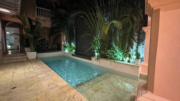 A swimming pool with tiled flooring, surrounded by potted plants and palm trees, with a building in the background.
