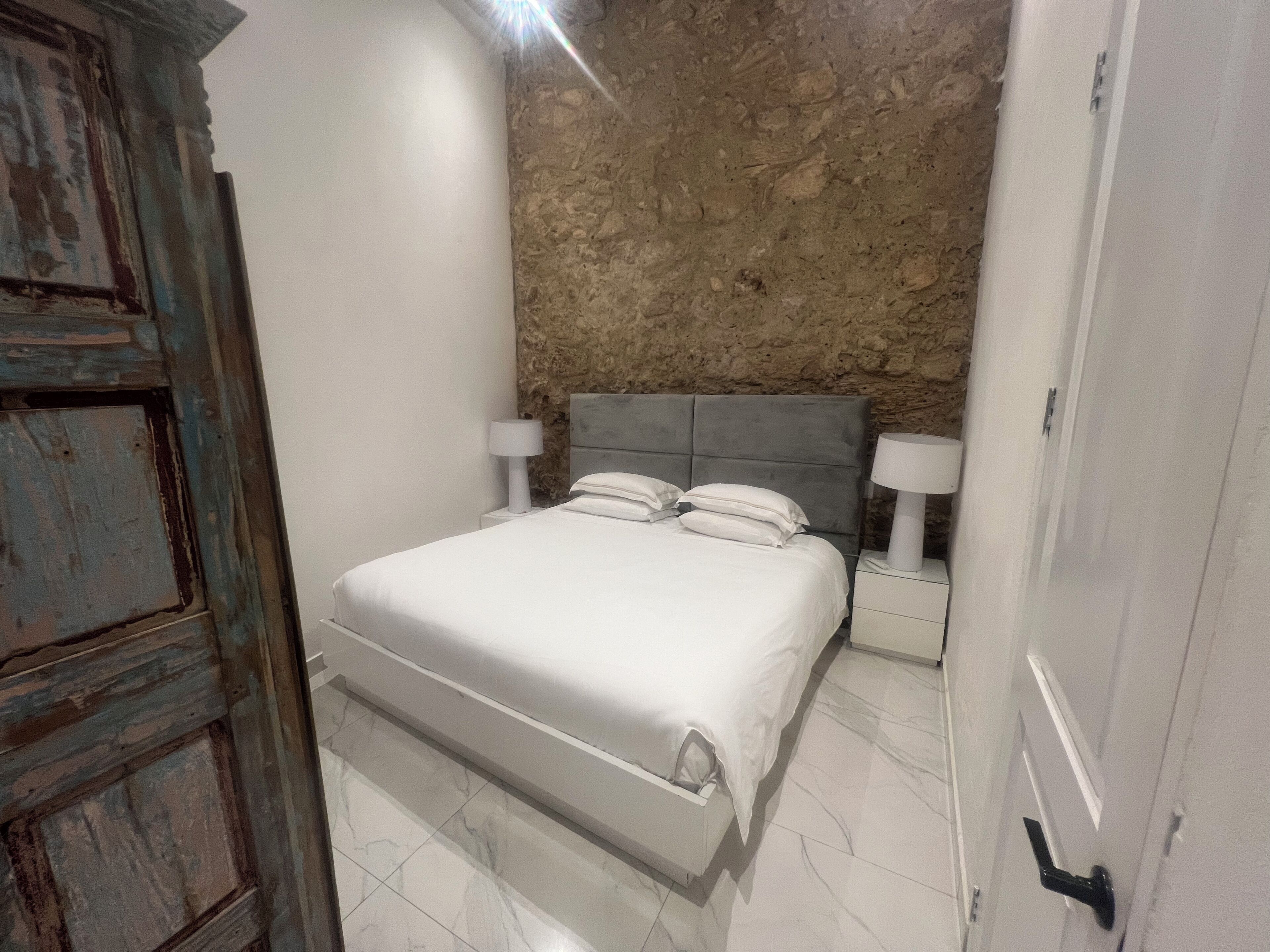 Un dormitorio con pared de piedra, una cama con ropa de cama blanca, dos mesitas de noche con lámparas y una puerta rústica.