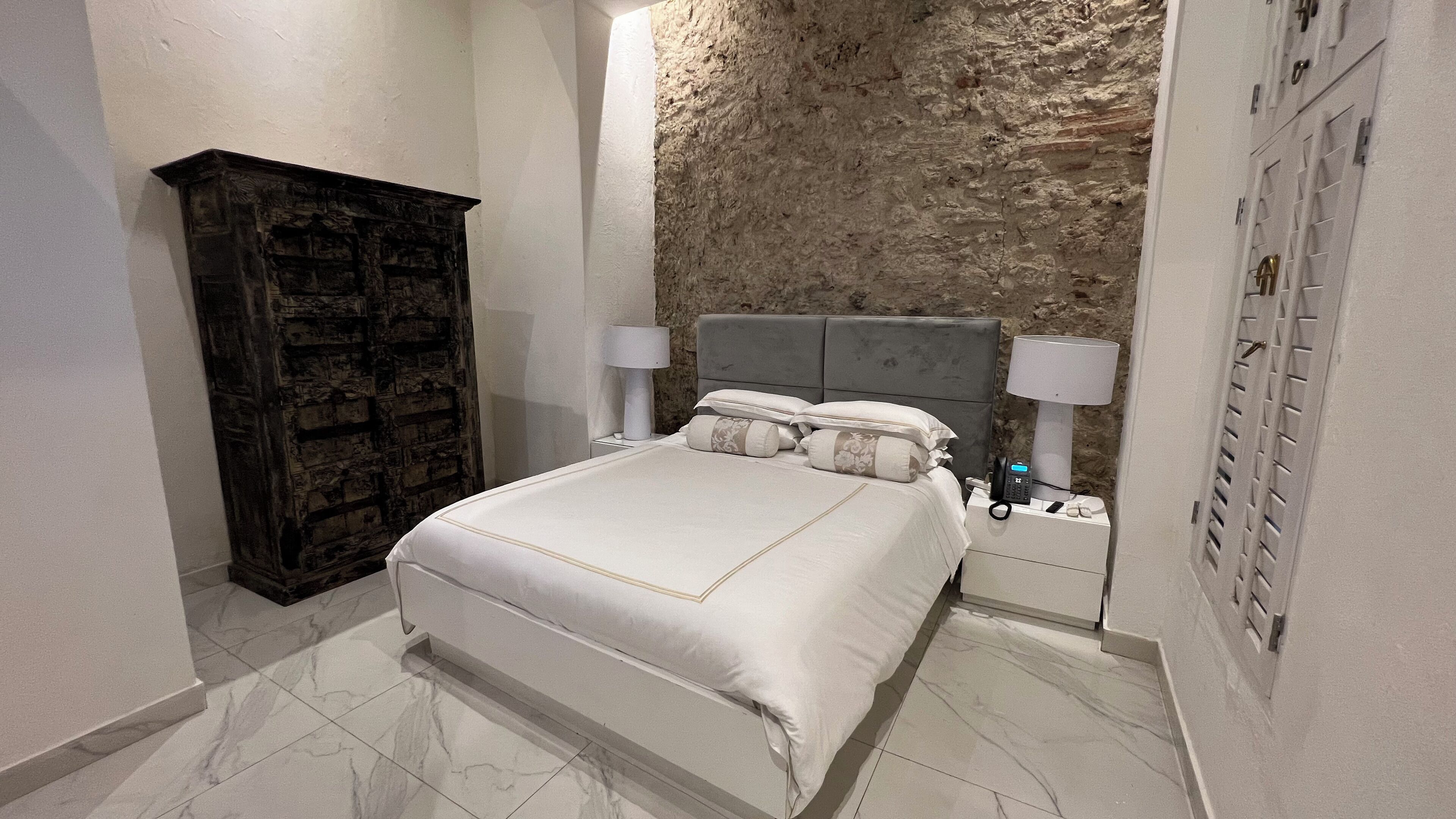 A bedroom with a bed, two bedside tables with lamps, a dark wooden wardrobe, and a textured stone wall.