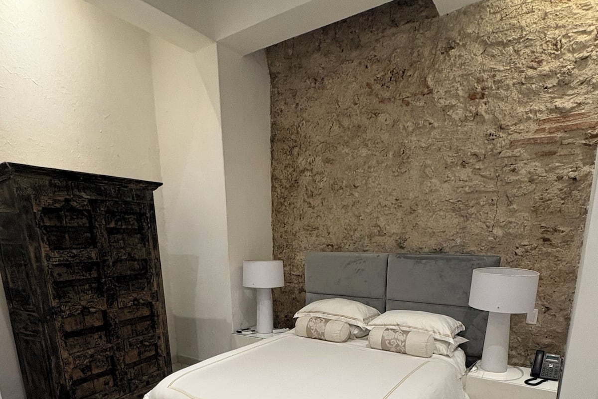 Quarto com piso de mármore, cama com roupa de cama branca, parede de pedra e guarda-roupa de madeira escura.