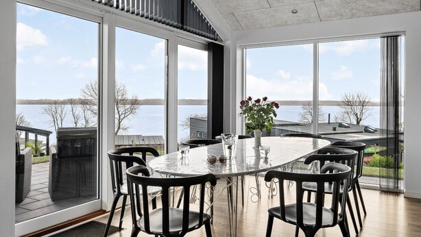 A dining area with a glass table and black chairs, large windows with a view of a lake and buildings, and a ceiling with a textured finish.