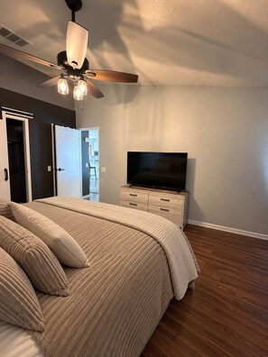 Un dormitorio con cama, televisor en una cómoda, ventilador de techo y puerta que da a otra habitación.