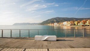 A waterfront promenade with a modern white bench, a metal railing, and a view of colorful buildings along the coast. - Nice home in Lusignano d'Albenga (Lusignano d'Albenga)