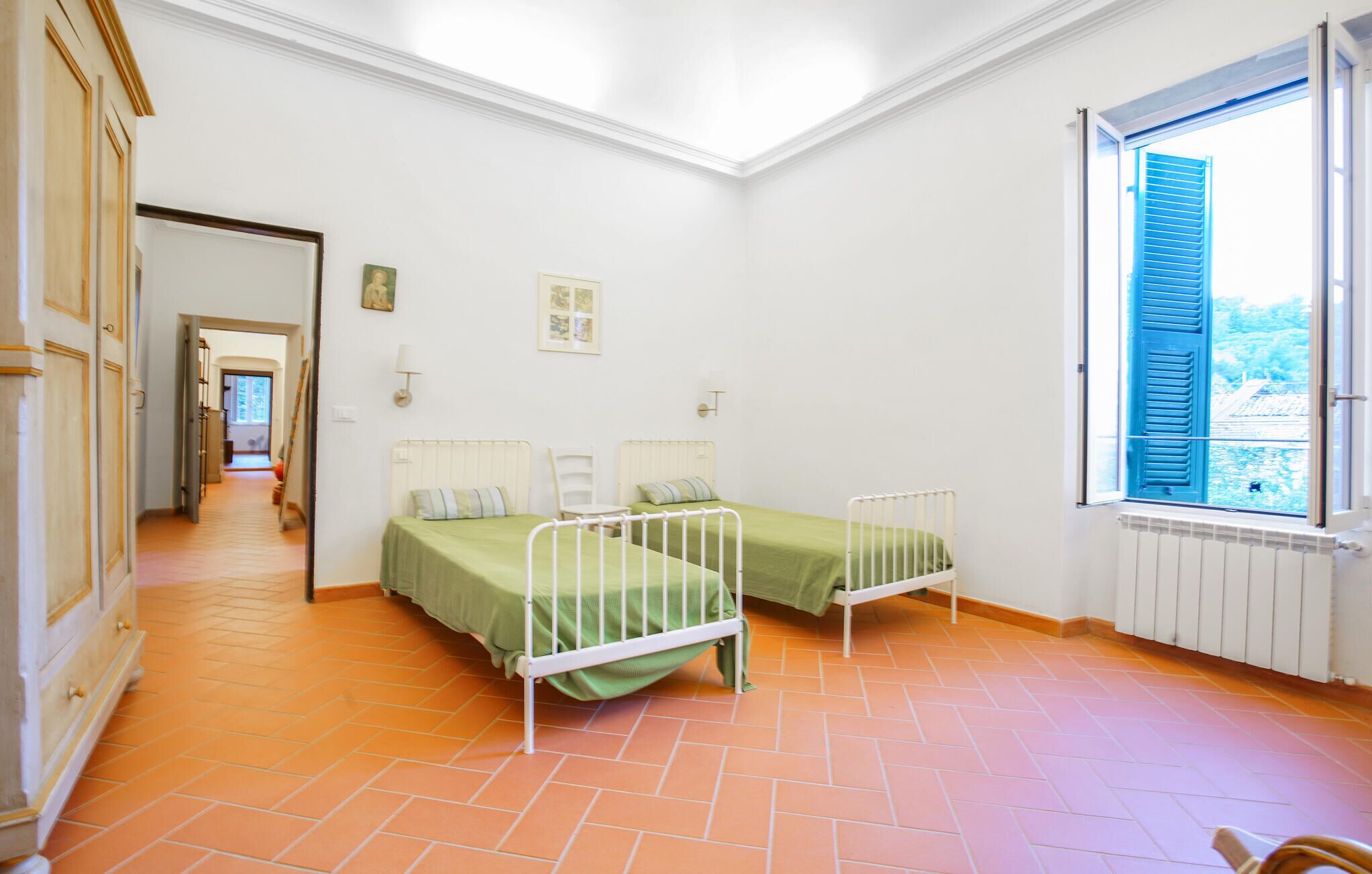 Ein Zimmer mit zwei Betten, einem Heizkörper, einem Fenster mit blauen Läden und einem zur anderen Kammer führenden Flur.