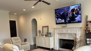 Een woonkamer met een open haard, witte leren banken, een flatscreen televisie en een plafondventilator.