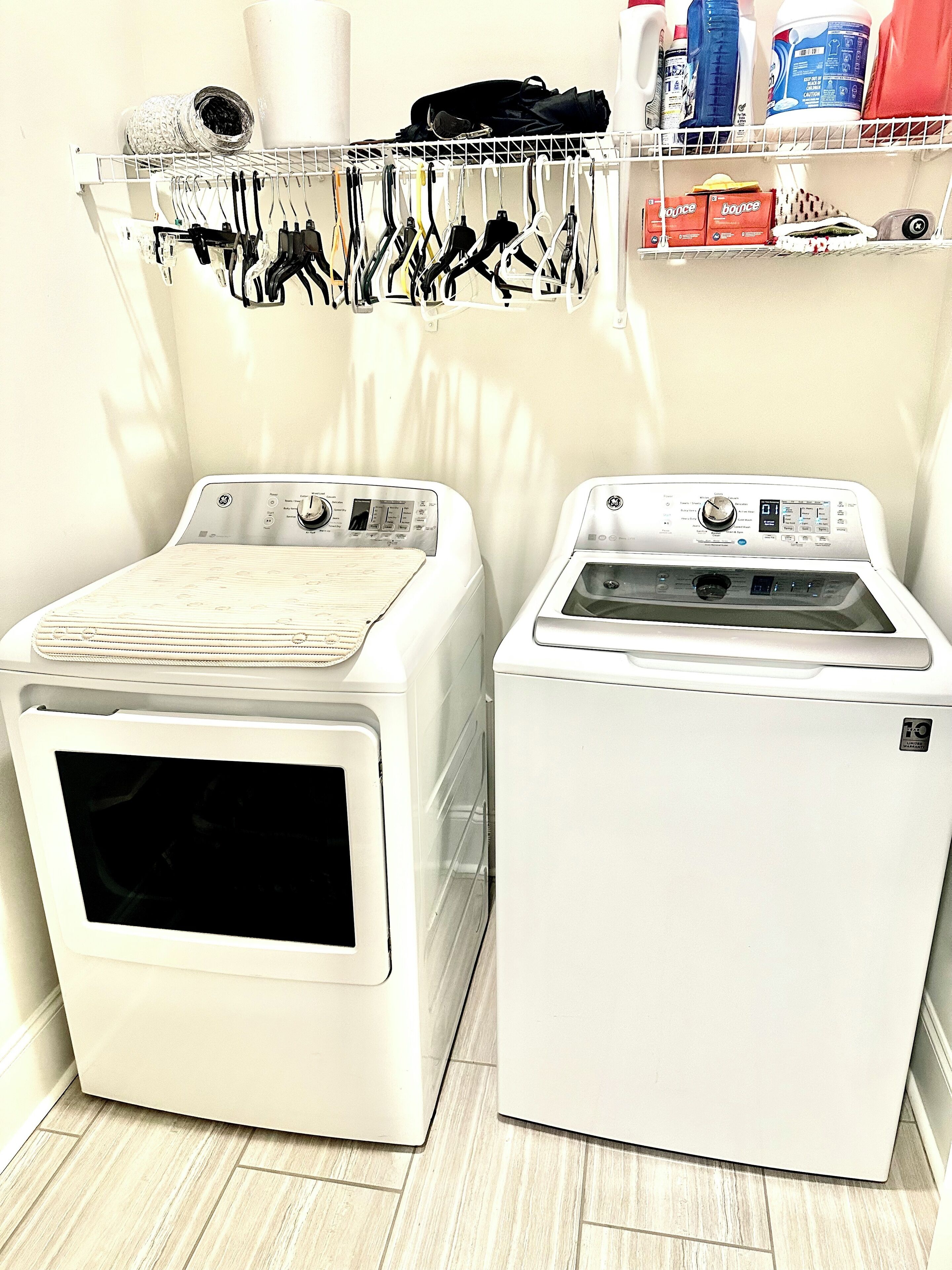 Two front-loading washing machines in a laundry room with a shelf above holding various cleaning supplies and a rack with hanging clothes.