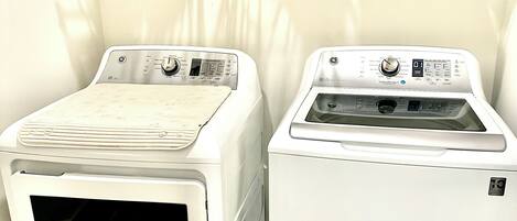 Two front-loading washing machines in a laundry room with a shelf above holding various cleaning supplies and a rack with hanging clothes.