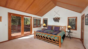 A bedroom with a wooden bed, two bedside lamps, a small table, and a view of a wooden deck through a sliding glass door. - Mountain Home with Private Hot Tub (Silverthorne)