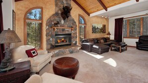 A cozy living room with a stone fireplace, leather furniture, and large windows offering a view of the outdoors. - Mountain Home with Private Hot Tub (Silverthorne)