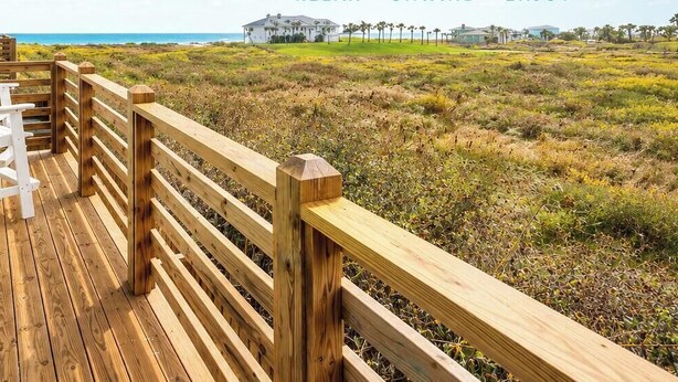 A wooden deck with white chairs and a wooden railing overlooking a beach.