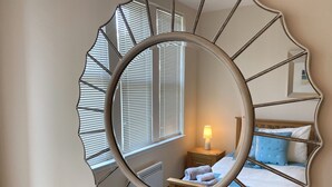 Ein Schlafzimmer mit großem Spiegel, einer Nachttischlampe, einem Bett mit blau-weißer Bettwäsche und einem Fenster mit Jalousien.