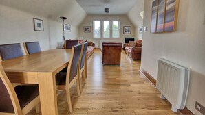 Ein geräumiges Wohnzimmer mit Esstisch aus Holz, gepolsterten Sitzmöglichkeiten und großen Fenstern.