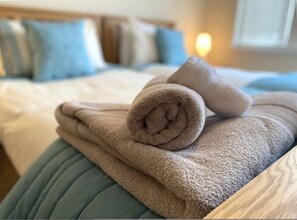 Letto rifatto con cura, un asciugamano beige piegato e un asciugamano arrotolato su una testiera imbottita blu.