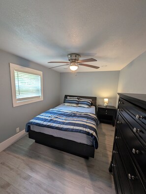 A bedroom with a bed, a ceiling fan, a nightstand, a dresser, and a window with blinds.