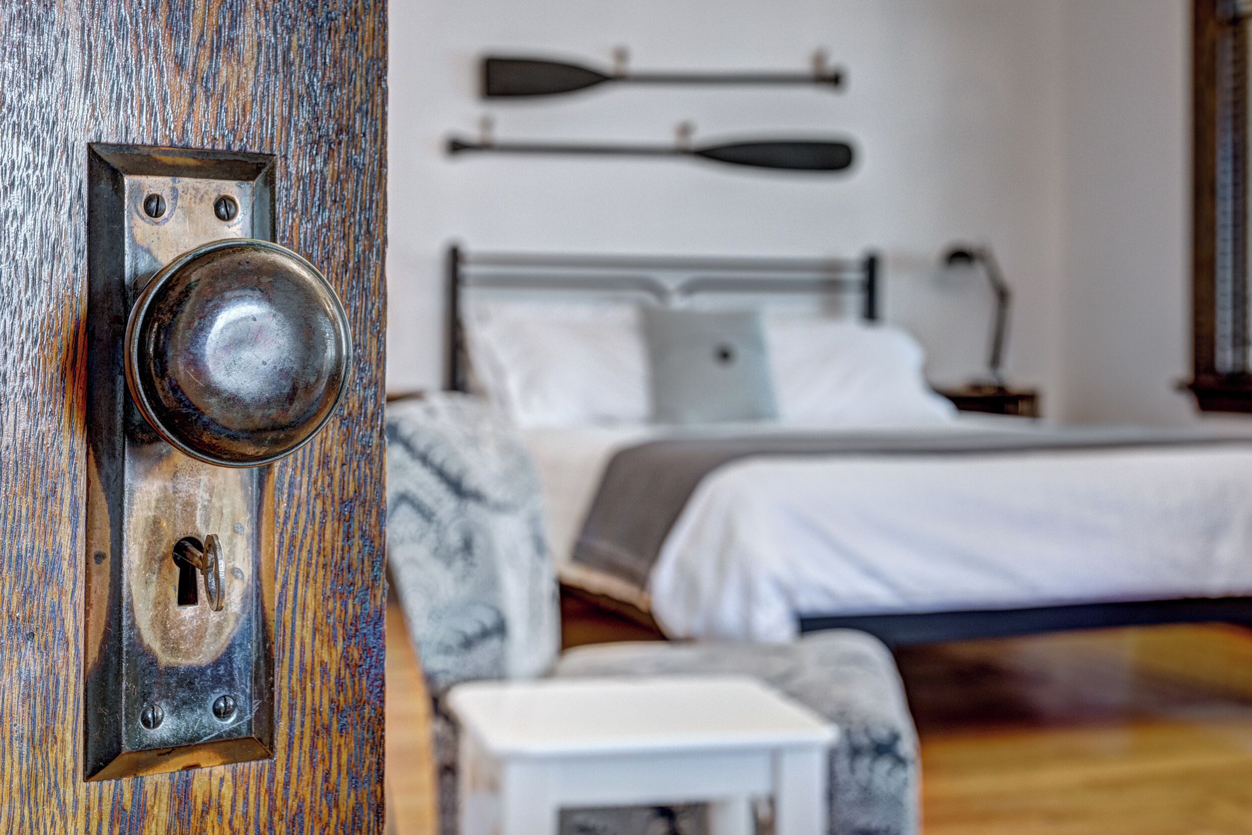 Eine Holztür mit Schlüsselloch und einem Metallgriff, ein Bett mit weißer Bettwäsche und ein Nachttisch mit einer Lampe.