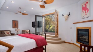 Un dormitorio con cama, escritorio, silla, televisión, ventilador de techo, chimenea y ventana con vistas a árboles.