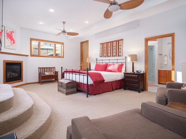 A spacious bedroom with a large bed, two bedside tables, a wooden chair, a fireplace, a ceiling fan, and a view of the outdoors through a window.