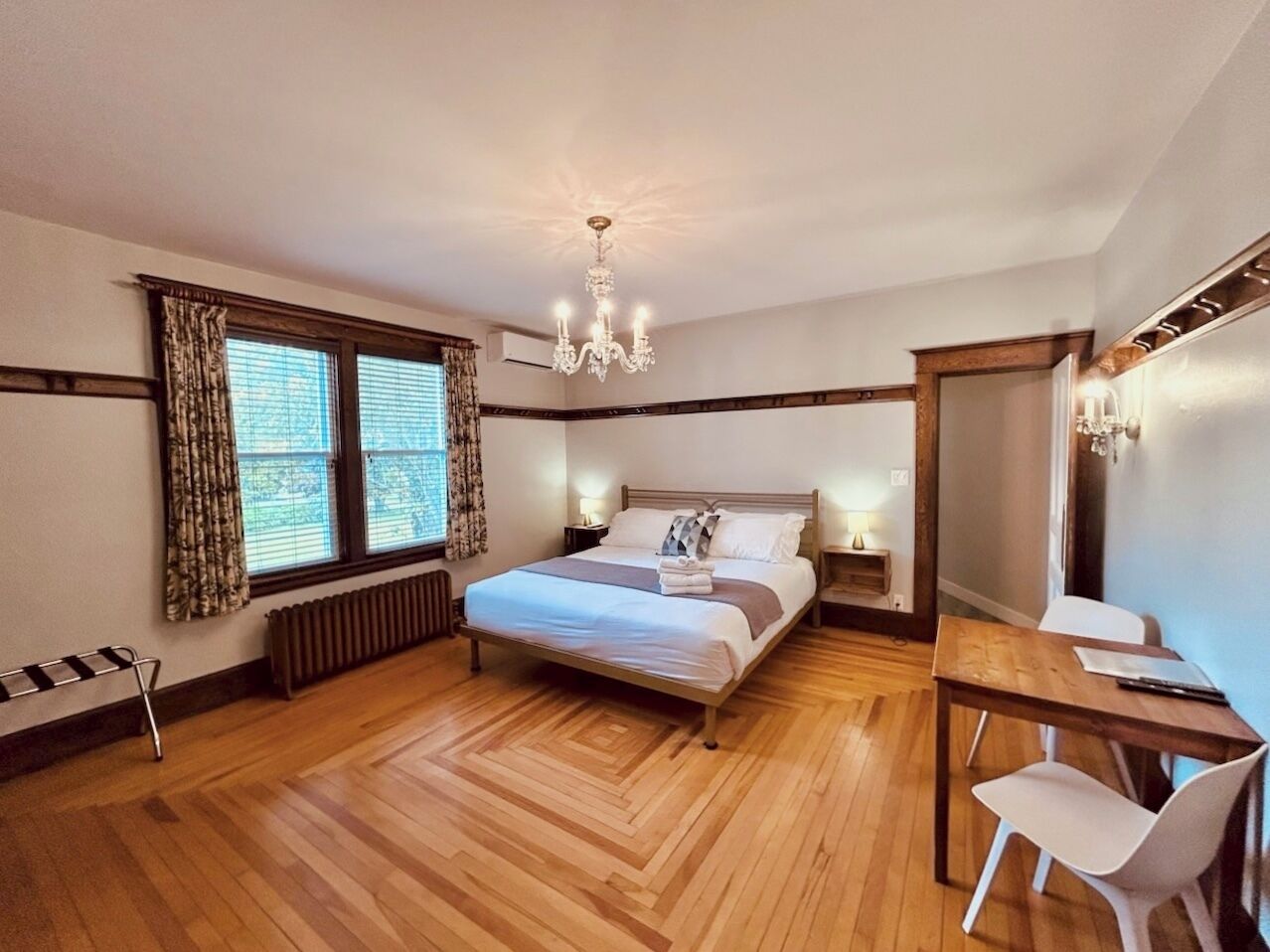 Une chambre à coucher comprenant un lit, des tables de chevet, un bureau, une chaise, un radiateur, une fenêtre avec des rideaux et un lustre.