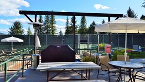 An outdoor patio with a grill, tables, and umbrellas, surrounded by a tennis court and trees. - RMR: Tensleep B4 Condo in Teton Village (Teton Village)