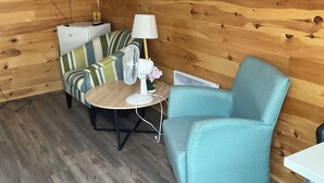 A cozy room with wooden walls, a flat-screen TV mounted on the wall, a striped sofa, a teal armchair, a small round table with a lamp, and a mirror.